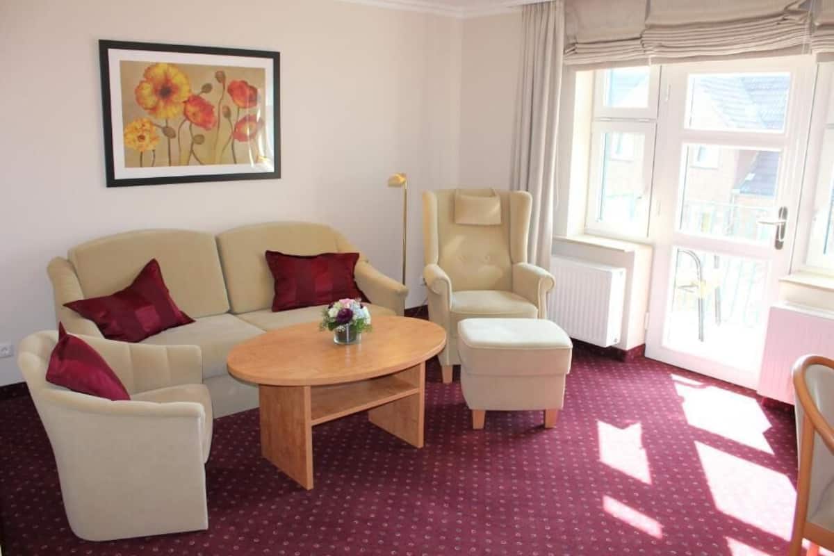 A living room with a beige sofa, two armchairs, a wooden coffee table, and a floor lamp. There is a window with curtains and a radiator.