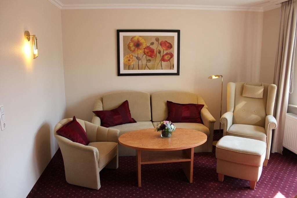 Ein Hotelzimmer mit einem beigefarbenen Sofa, zwei Sesseln, einem Holztisch und einem gerahmten Blumenbild an der Wand.