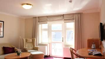 A hotel room with a sofa, armchair, ottoman, and a small table with a lamp. There is a door leading to the outside.