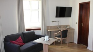Une pièce avec un canapé noir, une petite table sur laquelle se trouve un plateau, un bureau avec une chaise, un téléviseur fixé au mur et une porte en bois.