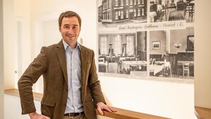 Un homme, vêtu d’un blazer marron et d’une chemise bleu clair, est appuyé contre une rambarde en bois. En arrière-plan, on aperçoit des photographies anciennes d’un bâtiment.