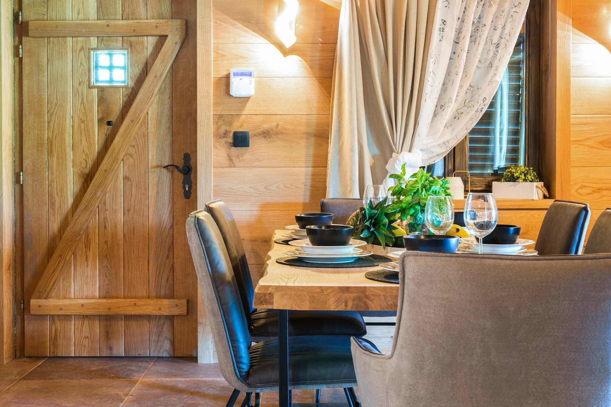 Een kamer met houten lambrisering, een eettafel gedekt voor twee personen, een raam met gordijnen en een thermostaat aan de muur.