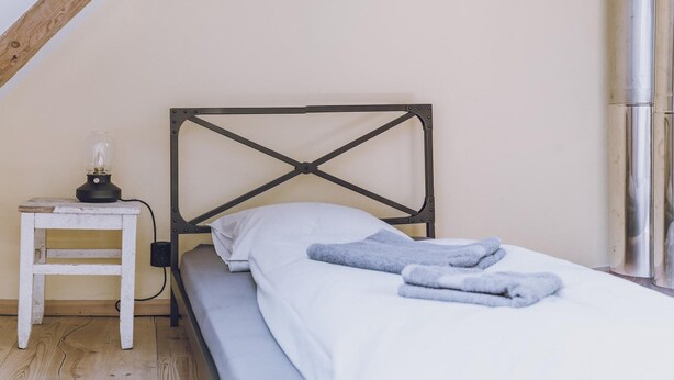 Una cama individual con cabecera de listones cruzados, una mesita de noche con lámpara y una ventana con cortina metálica.