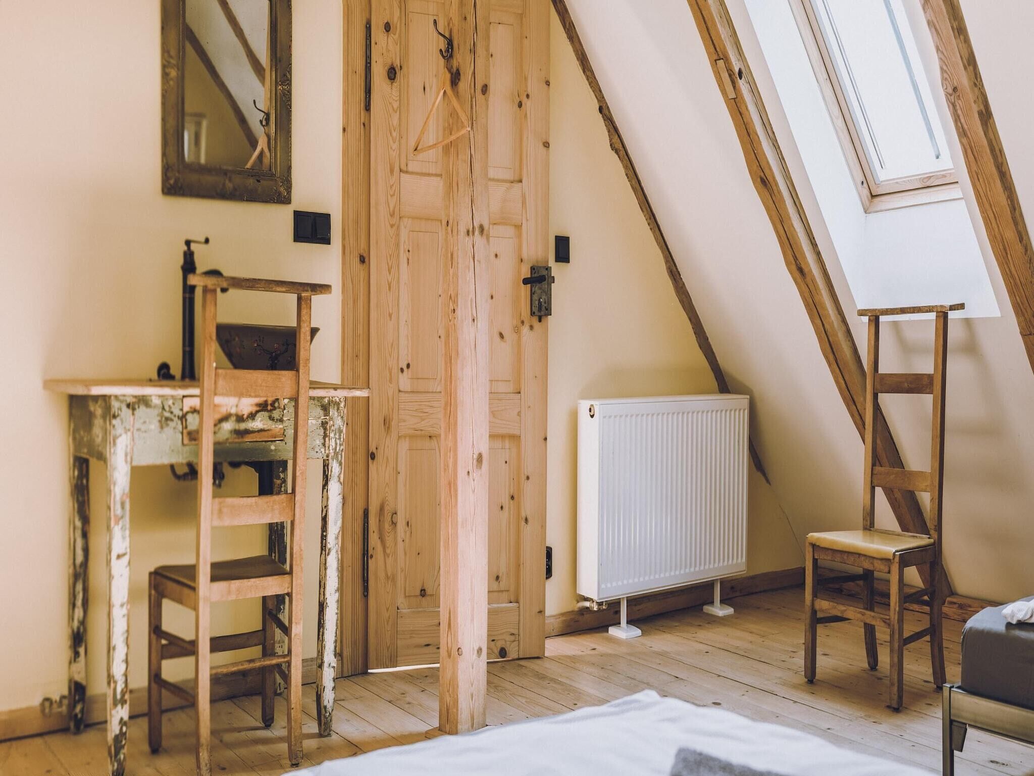 A room with a sloped ceiling, a wooden door, a radiator, a desk, a chair, and a ladder.