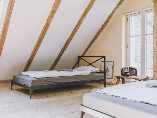 A bedroom with a sloped ceiling, a bed, a small table with a radio, and a window with a view of trees.