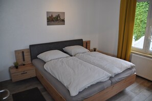 Un lit double avec une tête de lit grise, une petite table de chevet ornée d’une plante et un tableau accroché au mur.