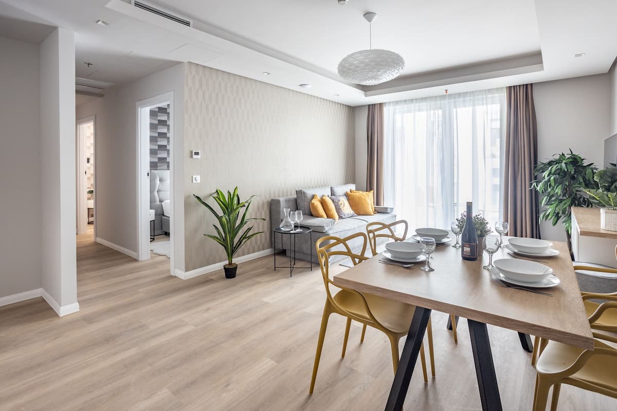 A modern dining area with a wooden table set for four, a grey sofa with yellow cushions, and a potted plant.