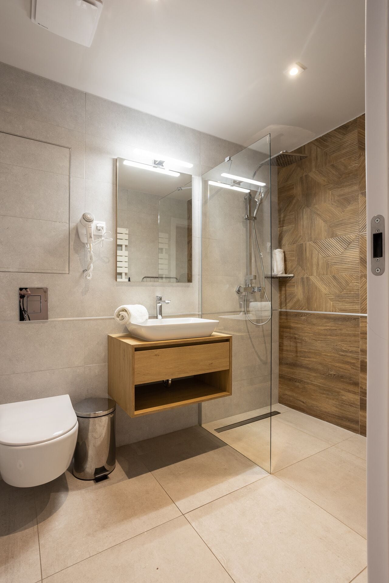 A modern bathroom with a glass-enclosed shower, a wooden vanity, a white toilet, and a trash can.
