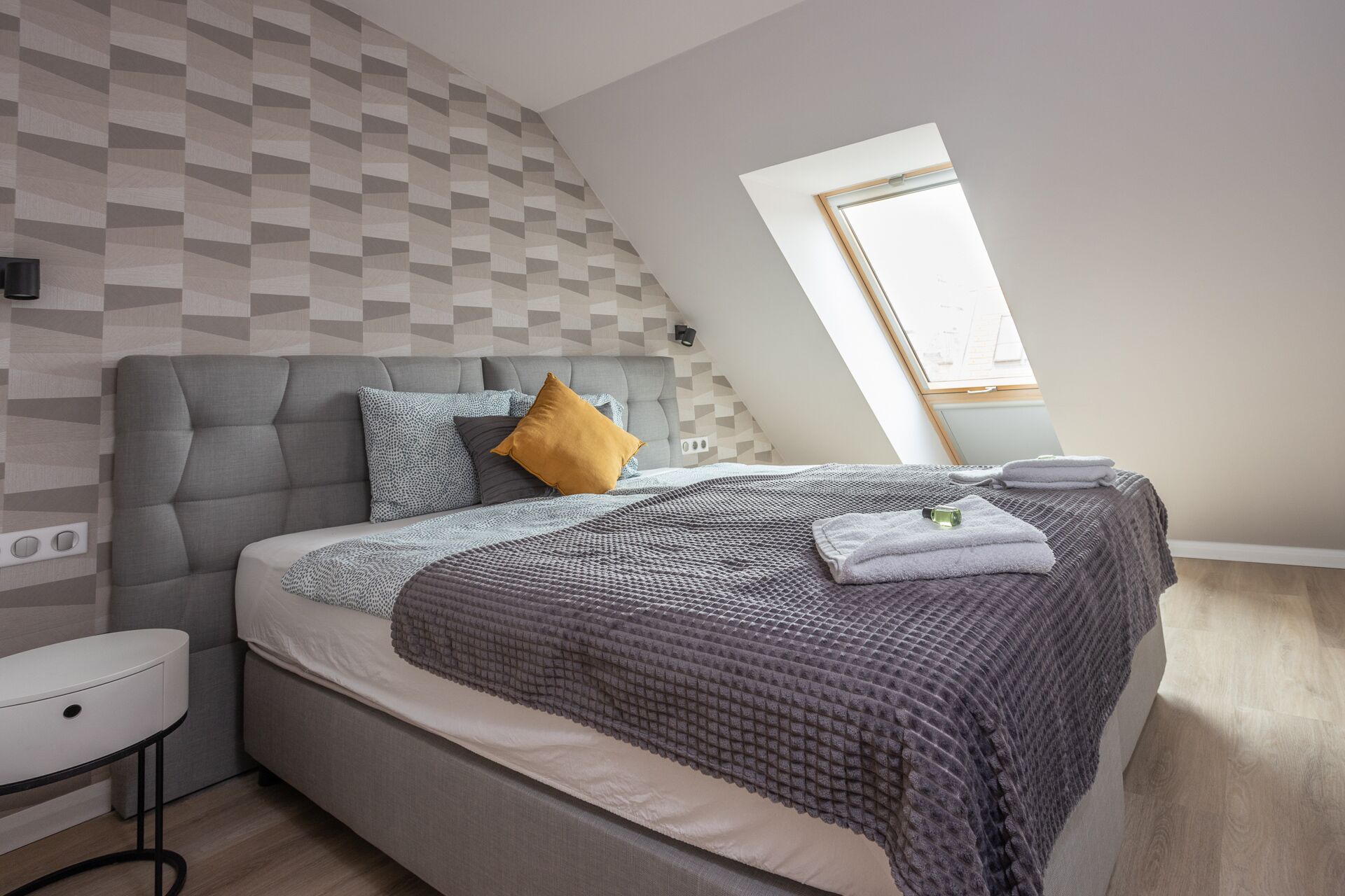 A modern bedroom with a loft-style ceiling, a grey upholstered headboard, a patterned wall, and a small bedside table.