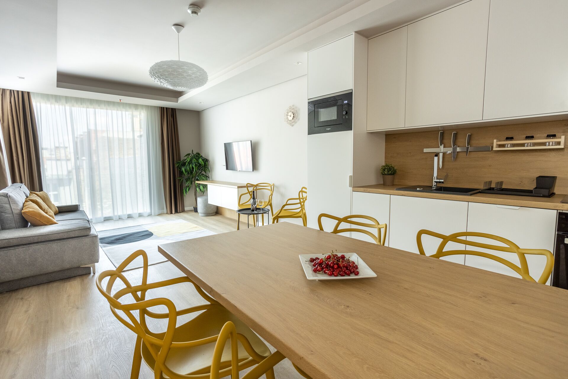 Une cuisine moderne avec des meubles blancs, un four à micro-ondes et un bloc à couteaux. On y trouve une table à manger avec des chaises jaunes et une assiette de cerises.