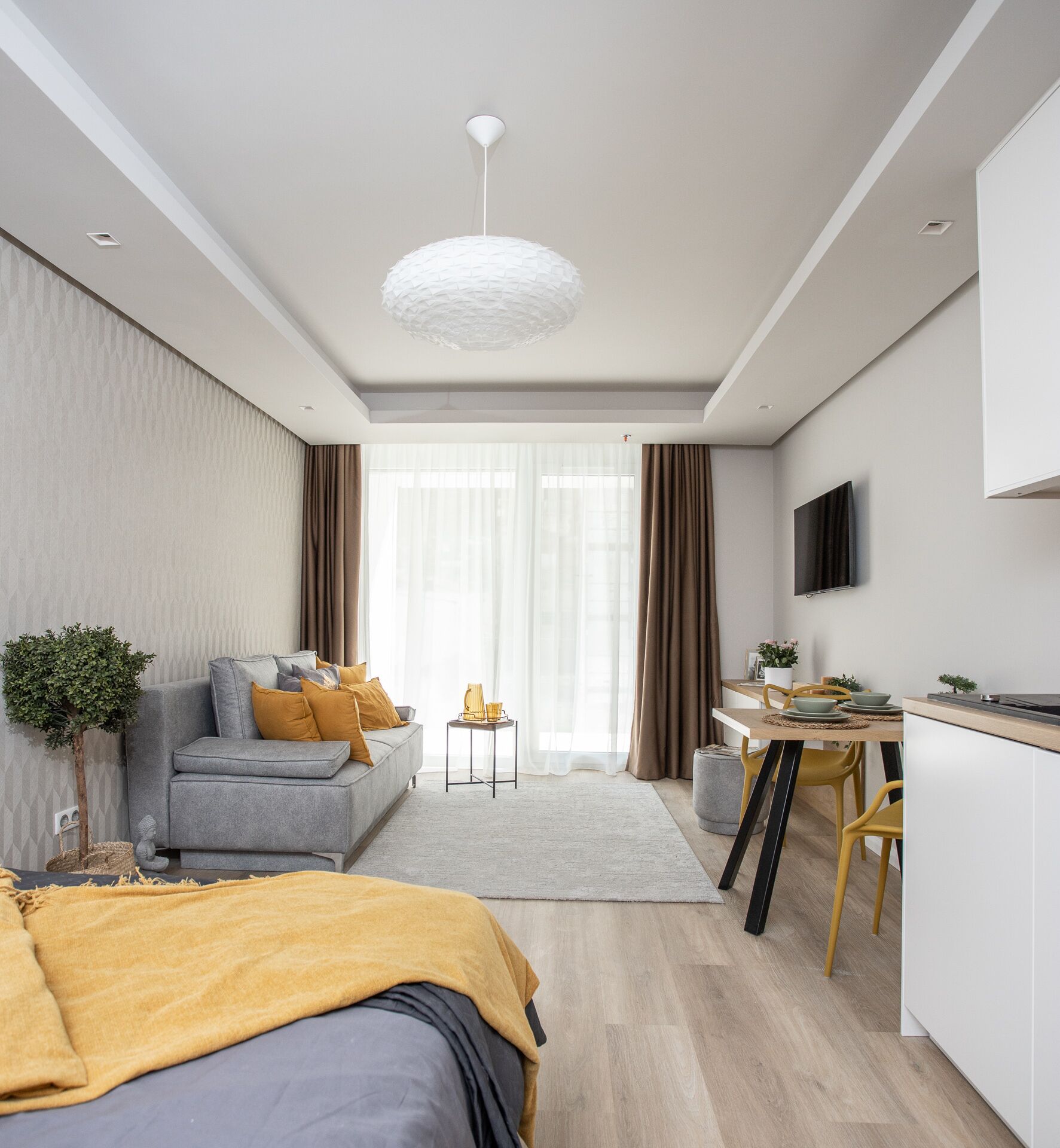 A modern, well-lit living room with a grey sofa, a dining area with a wooden table, and a kitchen area with a TV mounted on the wall.