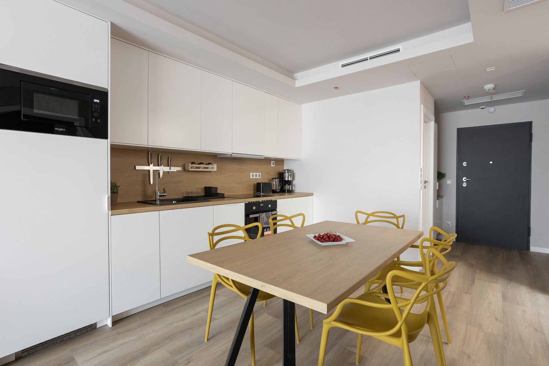 A modern kitchen with white cabinets, a built-in microwave, and a coffee machine. There is a wooden dining table with yellow chairs and a plate of cherries.