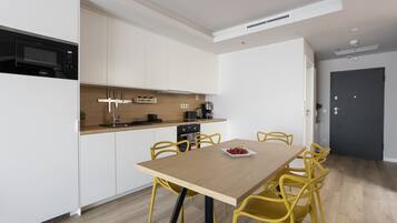 A modern kitchen with white cabinets, a built-in microwave, and a coffee machine. There is a wooden dining table with yellow chairs and a plate of cherries.