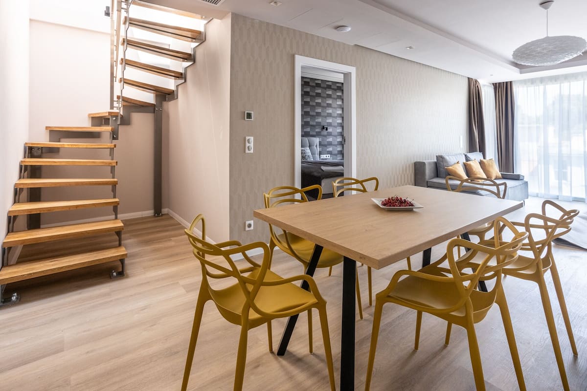 A modern living room with a wooden staircase, a dining area with yellow chairs, and a sofa with cushions.
