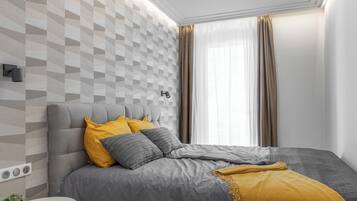 A modern bedroom with a grey sofa bed, a bedside table with a plant, and a large window with curtains.