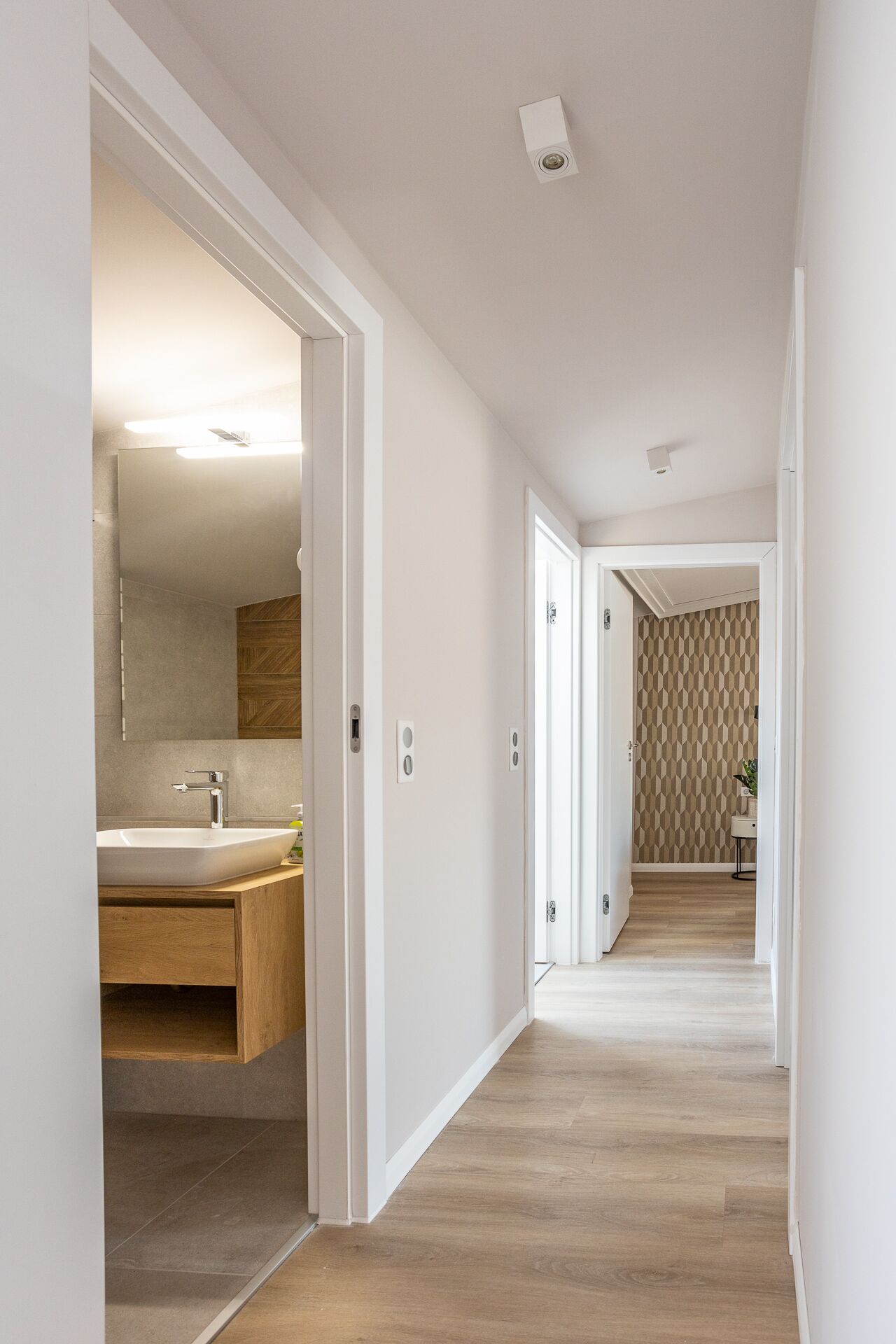 Un couloir moderne avec un plancher en bois, des murs blancs et une salle de bain équipée d’un lavabo et d’un miroir.