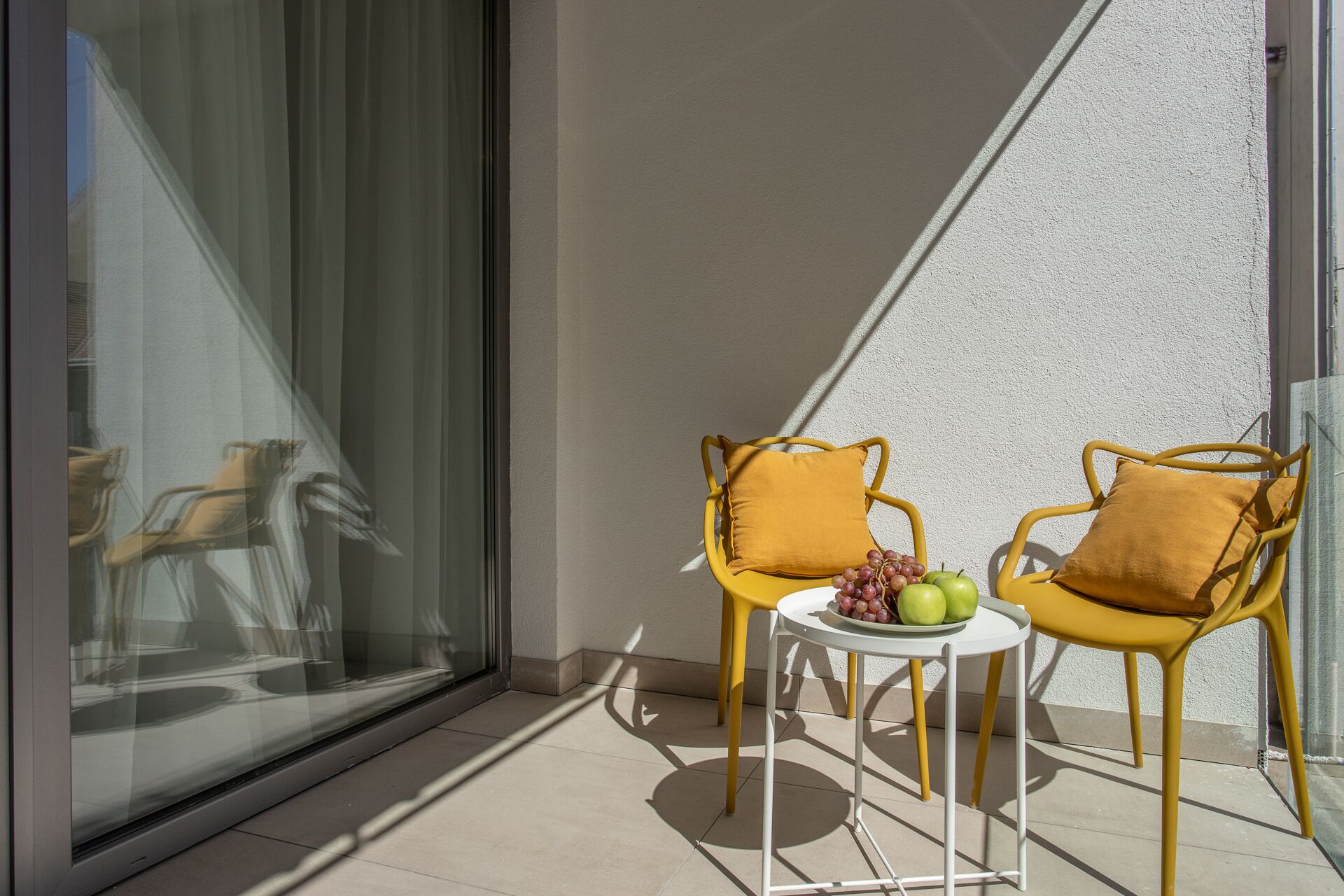A balcony with two yellow chairs and a small table, each with a cushion. A plate of green apples and grapes is on the table.