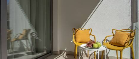 A balcony with two yellow chairs and a small table, each with a cushion. A plate of green apples and grapes is on the table.