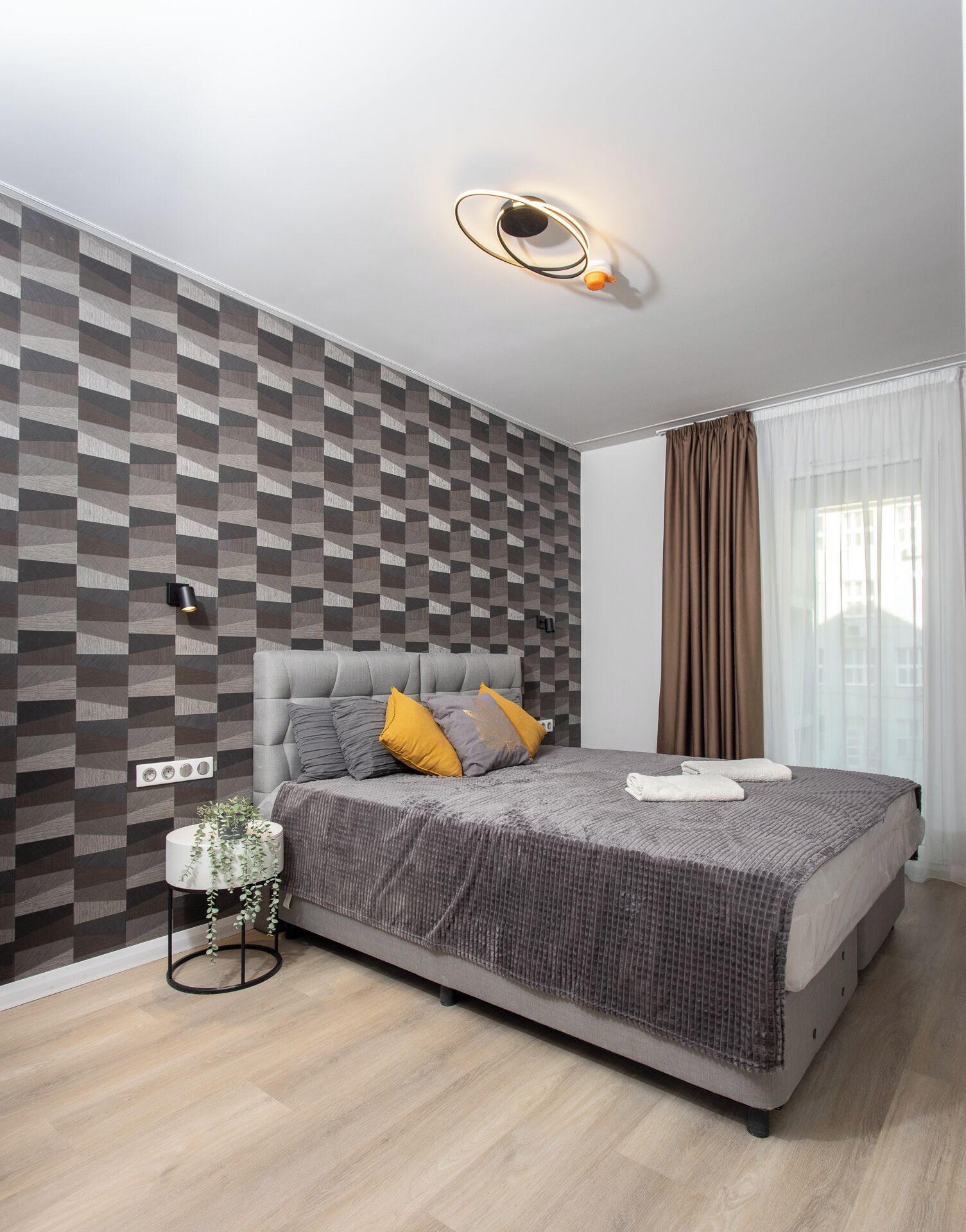 Ein modernes Schlafzimmer mit einer Wand im geometrischen Muster, einem gepolsterten Kopfteil und einem Nachttisch mit Pflanze.
