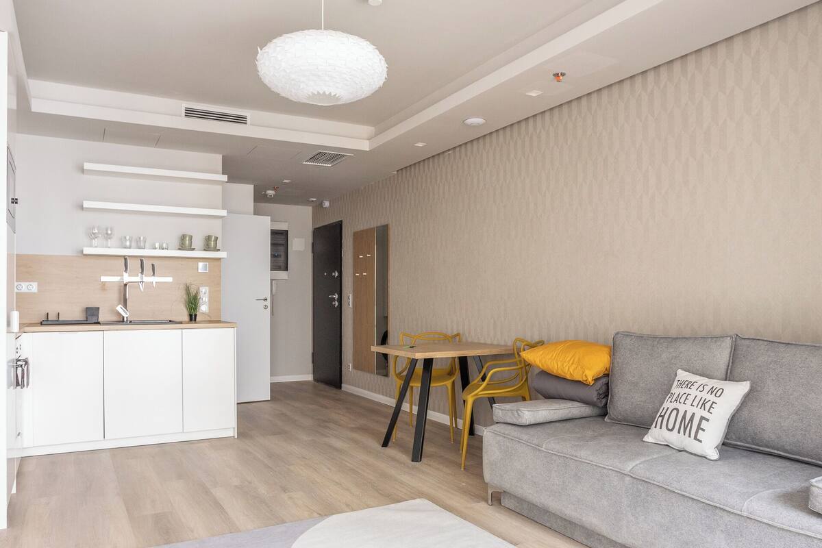 A modern living room with a grey sofa, a dining table with yellow chairs, and a kitchen area with white cabinets.