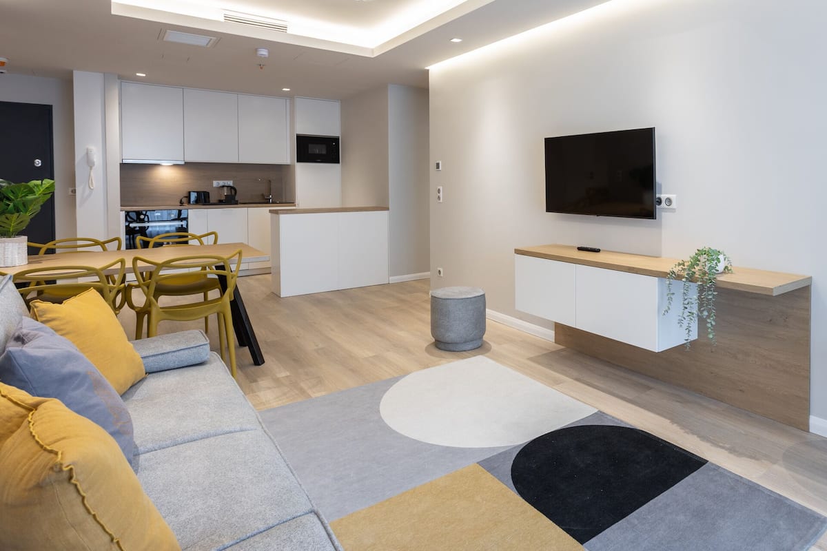 A modern living room with a sofa, a dining area, a kitchen, and a TV mounted on the wall.