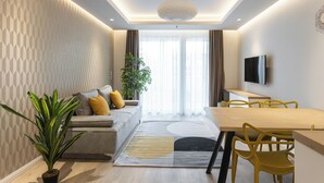 Un salon moderne comprenant un canapé, une table à manger avec des chaises, un téléviseur fixé au mur et une grande fenêtre avec des rideaux.