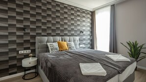Une chambre moderne avec un mur orné d’un motif géométrique, un lit recouvert d’un tissu gris, deux oreillers, une table de chevet avec une lampe et une plante en pot.