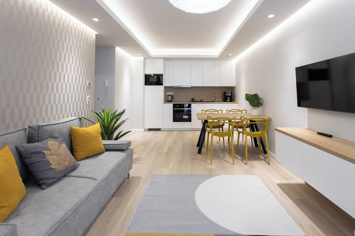A modern living room with a grey sofa, yellow cushions, a dining area with yellow chairs, and a kitchen with white cabinets and a TV mounted on the wall.