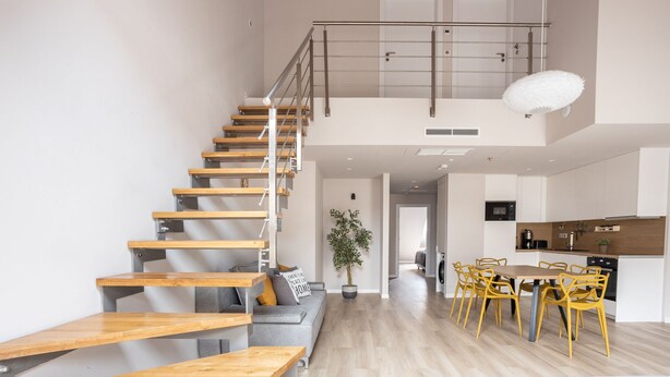 Un vaste espace de vie agrémenté d’un escalier en bois, un coin repas avec des chaises jaunes et une cuisine dotée de meubles blancs.