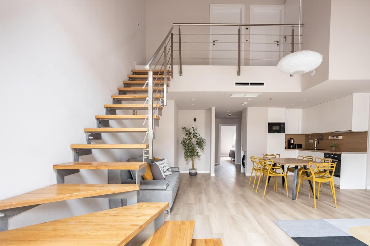A spacious living area with a wooden staircase, a dining area with yellow chairs, and a kitchen with white cabinets.