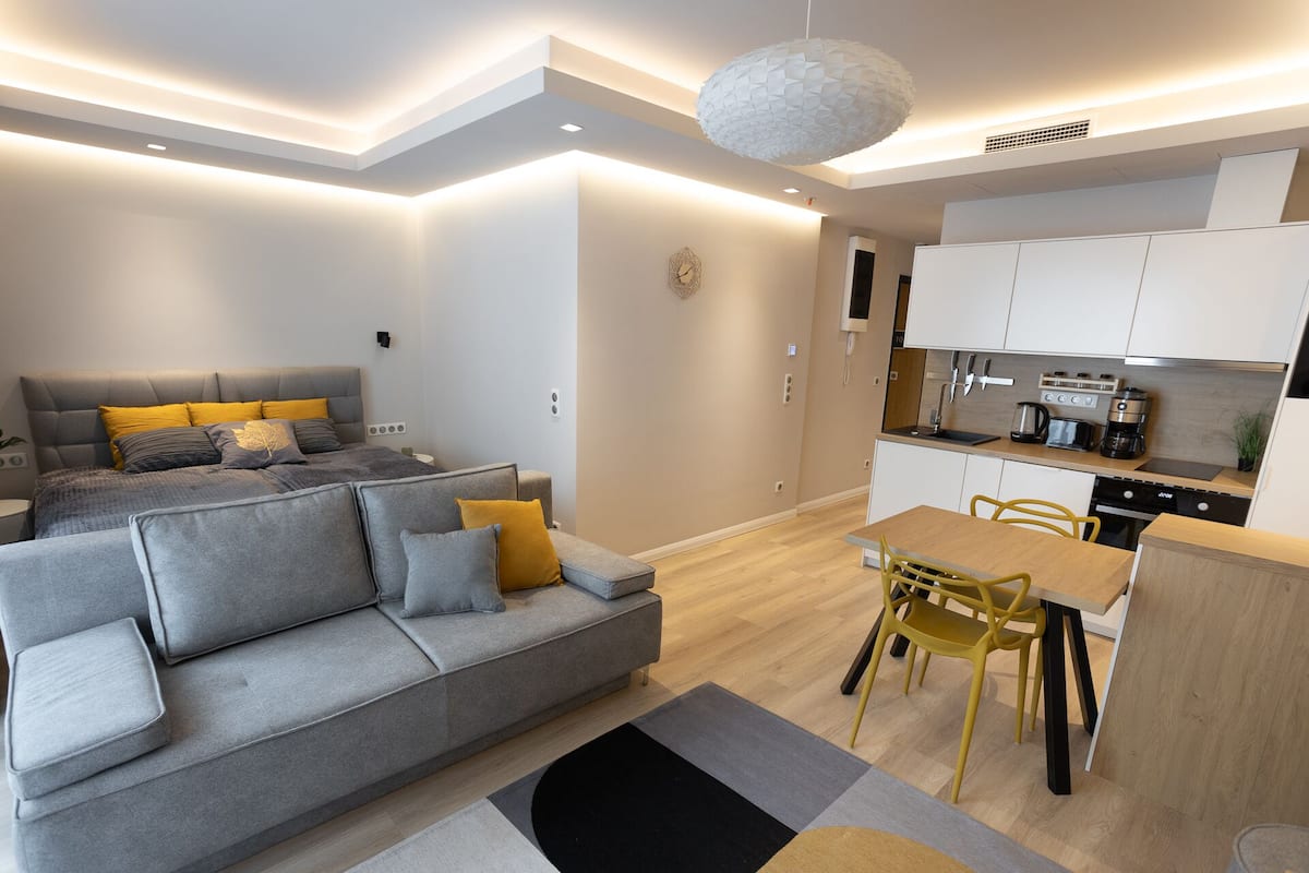 A modern apartment with a grey sofa, yellow cushions, a dining table with yellow chairs, and a kitchen area.