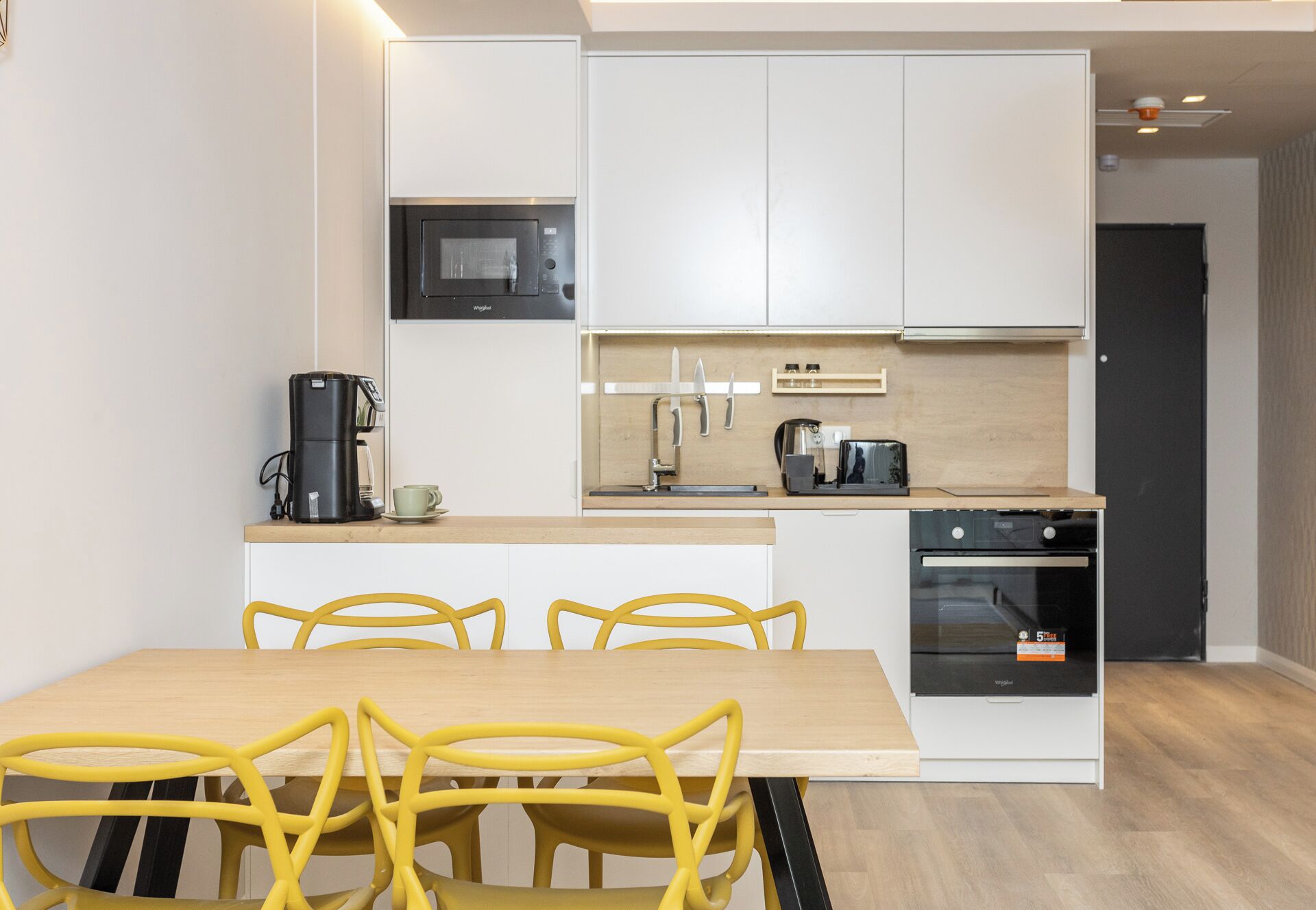 A modern kitchen with white cabinets, a microwave, a coffee maker, and a toaster. There is a wooden dining table with yellow chairs.