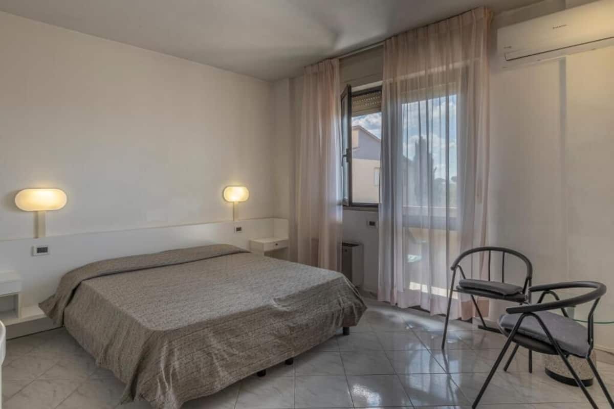Ein Schlafzimmer mit einem Bett, zwei Nachttischlampen, einem Sessel, einem Fenster mit Vorhängen und einer Klimaanlage.