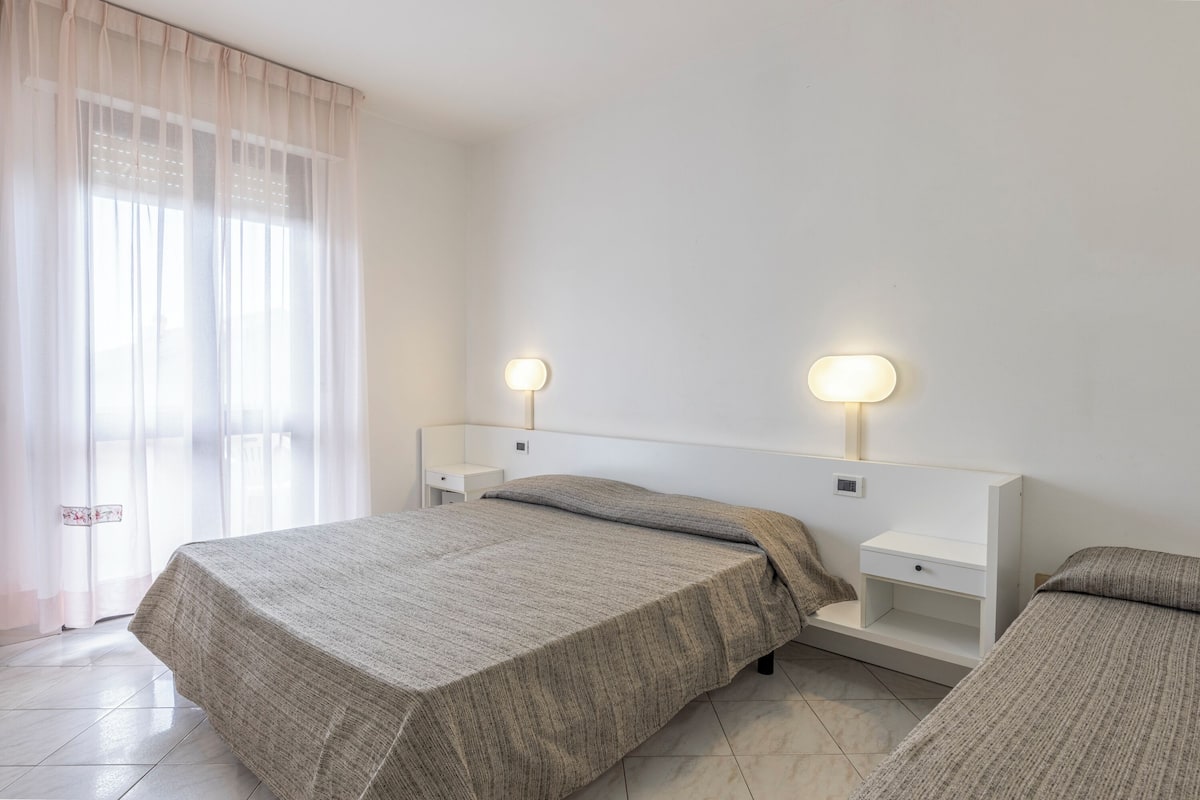 Ein Hotelzimmer mit Bett, Nachttischen und einem Fenster mit durchsichtigen Vorhängen.