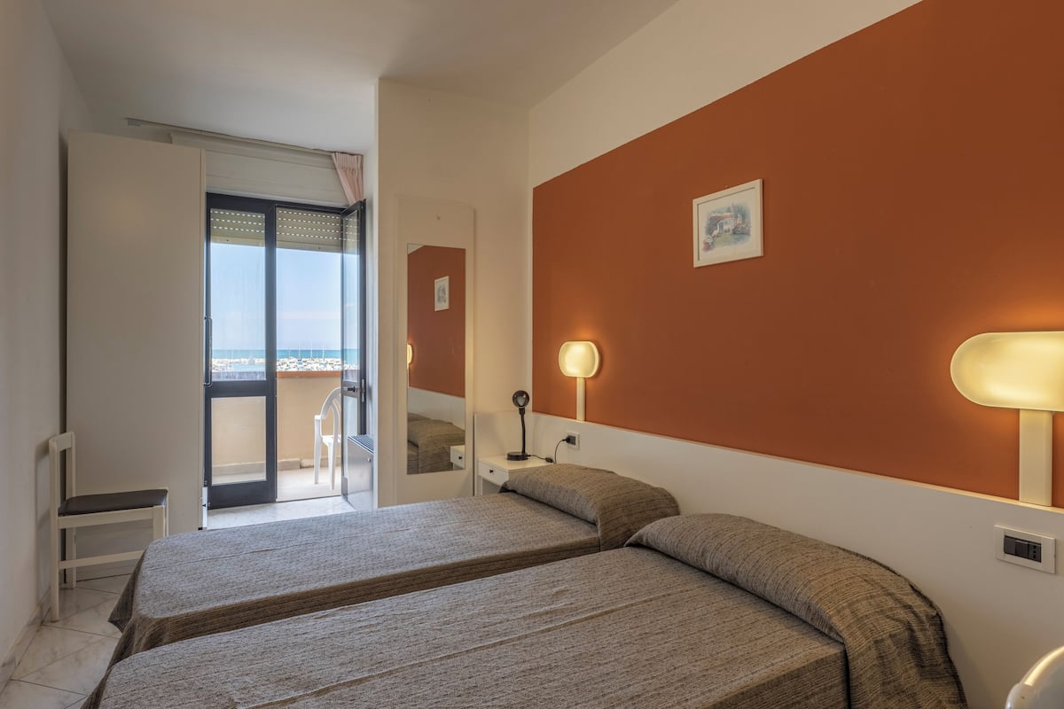 Ein Hotelzimmer mit zwei Betten, einem Schreibtisch, einem Sessel und einem Balkon mit Blick, sowie einer Wandlampe.