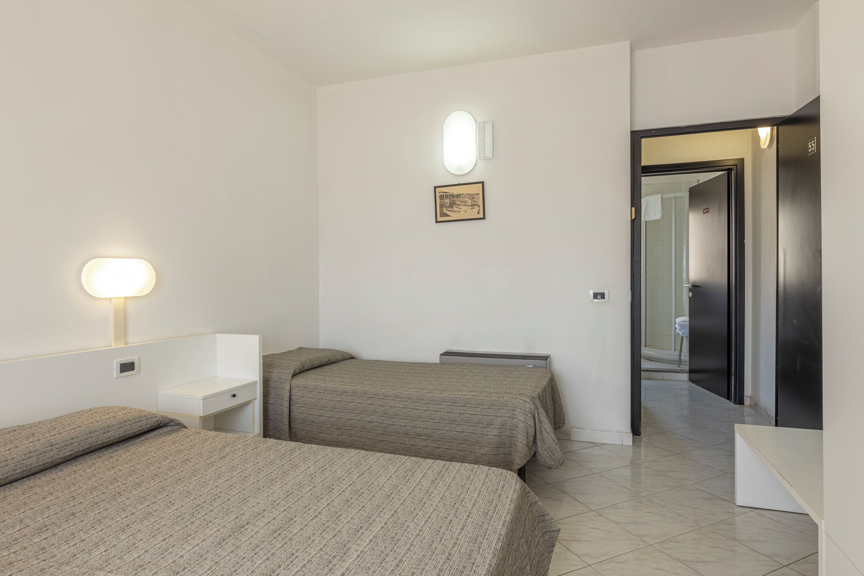 A hotel room with two beds, a desk, a wall-mounted light, and a framed picture.