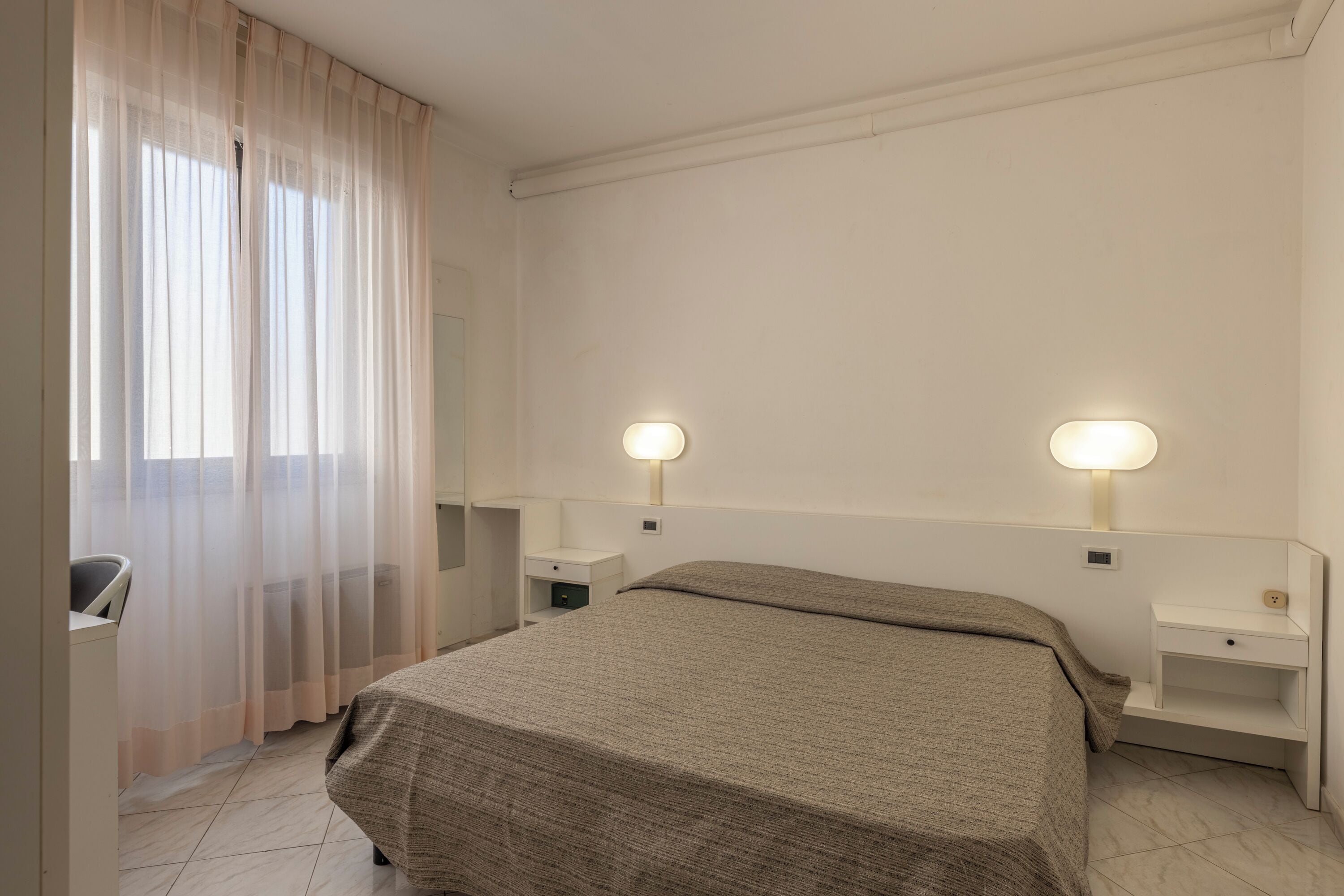 Une chambre d’hôtel avec un lit, deux tables de chevet, une chaise et une fenêtre avec des rideaux légers.