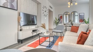 Un salon moderne doté d’un téléviseur à écran plat, d’un meuble de rangement blanc, d’une table basse ronde, d’un canapé gris avec des coussins orange et d’un tapis à motifs.