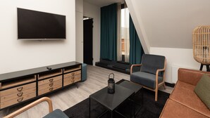 Een moderne woonkamer met een flatscreen televisie, een houten kast met lades, een zwarte salontafel, een blauwe fauteuil, een bruine bank en een rieten stoel.