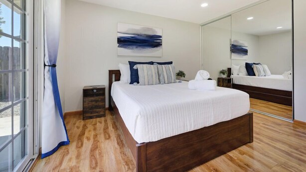 A modern bedroom with a large bed, a nightstand, a mirror, and a view through a sliding glass door.