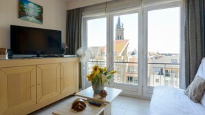 A hotel room with a large window, a wooden cabinet, a flat-screen TV, a small table with a vase of flowers, and a bed. - Simplistic Apartment in Blankenberghe near Belgium Pier (Blankenberghe)