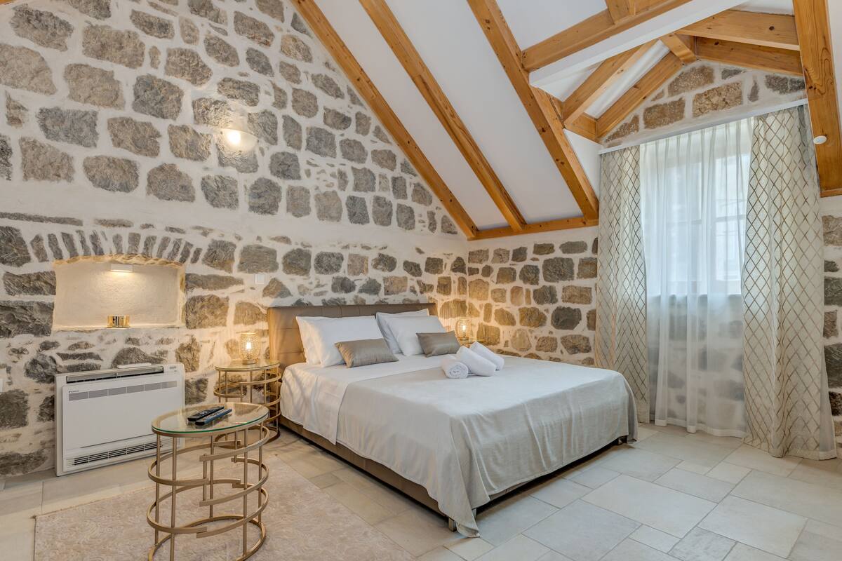 A bedroom with stone walls, a wooden ceiling, a bed with white bedding, a nightstand with a lamp, and a window with sheer curtains.