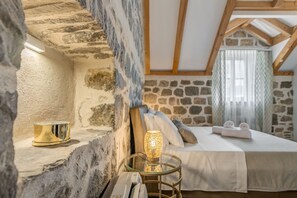 Una habitación acogedora con paredes de piedra, una cama con sábanas blancas, una mesa de vidrio con una lámpara y un cuenco dorado en una estantería.