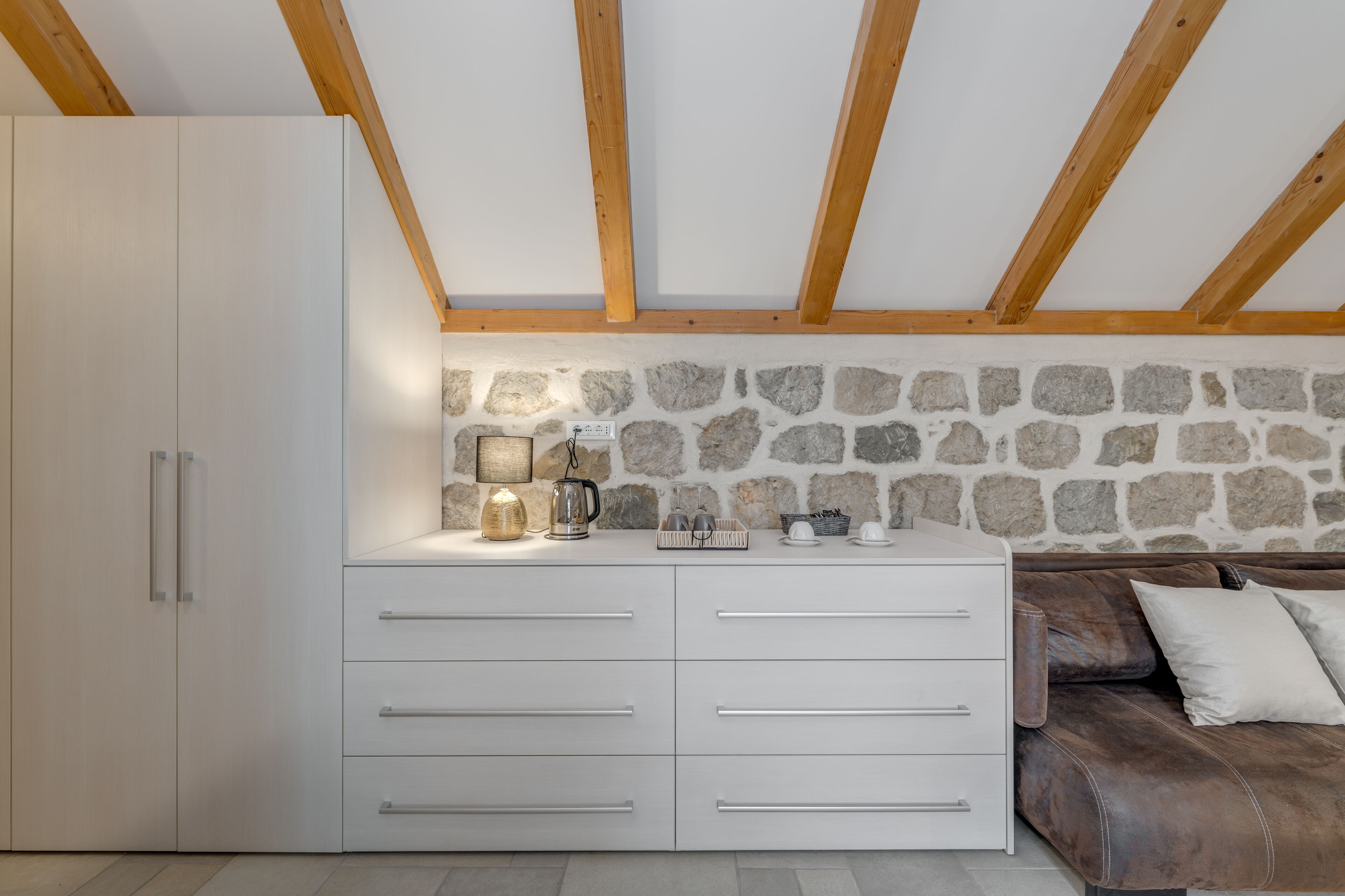 A modern kitchen with white cabinetry, a stone wall, and a wooden ceiling.