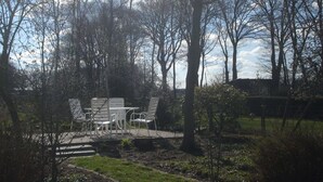 An outdoor seating area with white metal chairs and tables, surrounded by trees and a grassy lawn.