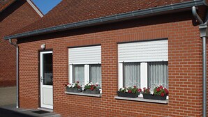 A brick building with two windows, each with a white blind and flower boxes containing red and white flowers.