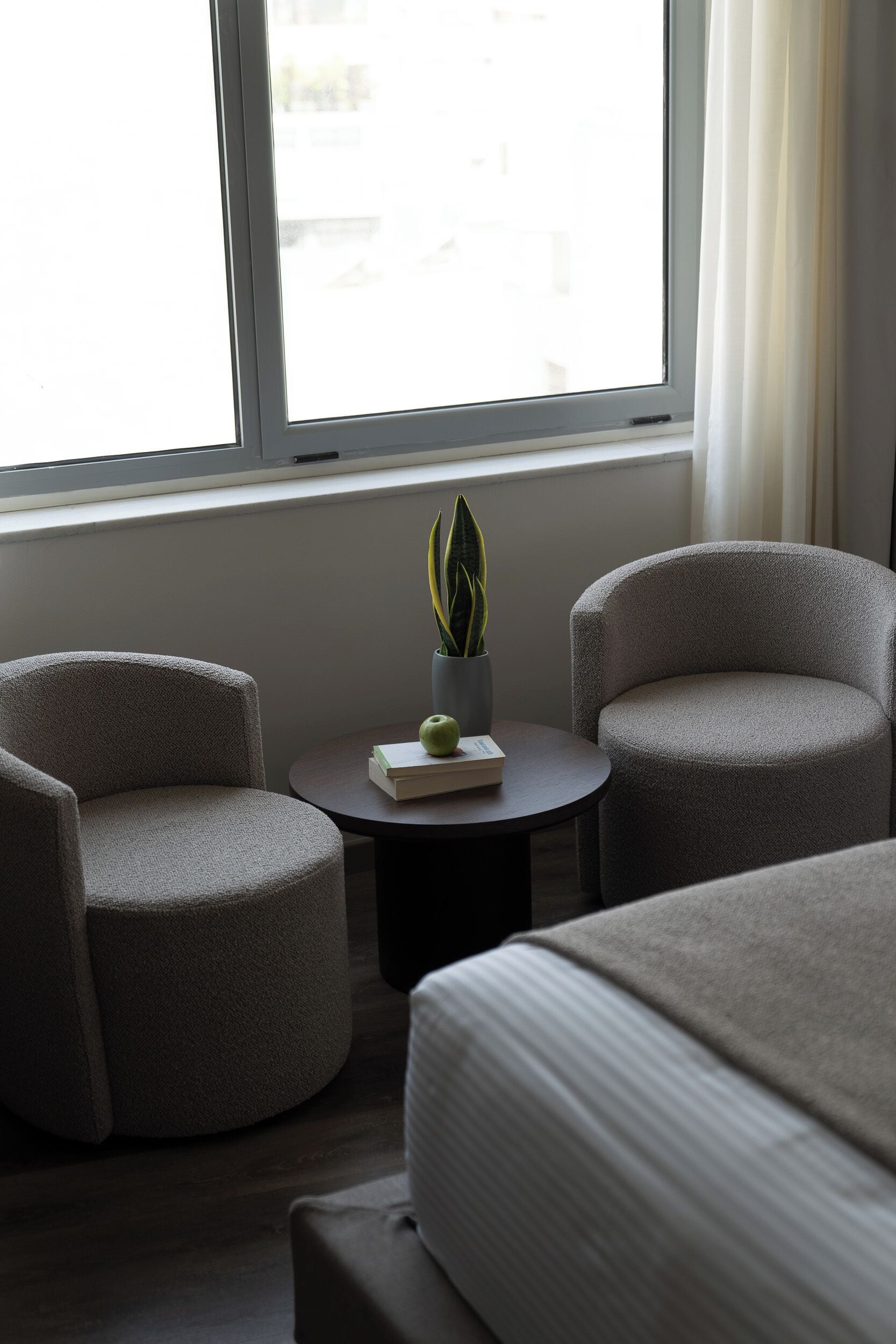 A modern hotel room with two armchairs, a small round table with a plant, and a bed with a striped bedspread.