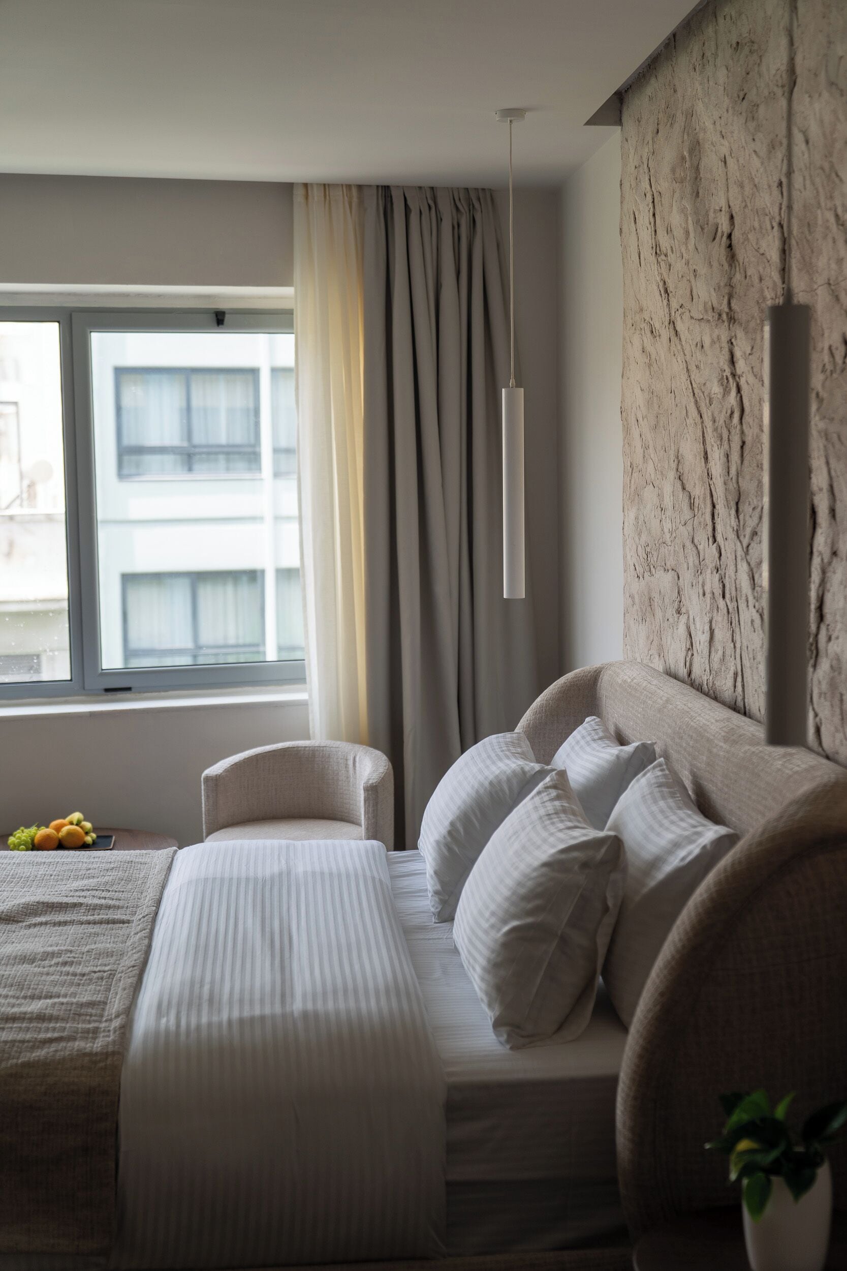 A modern bedroom with a large bed, a side table with a plant, and a view of the city through a window.