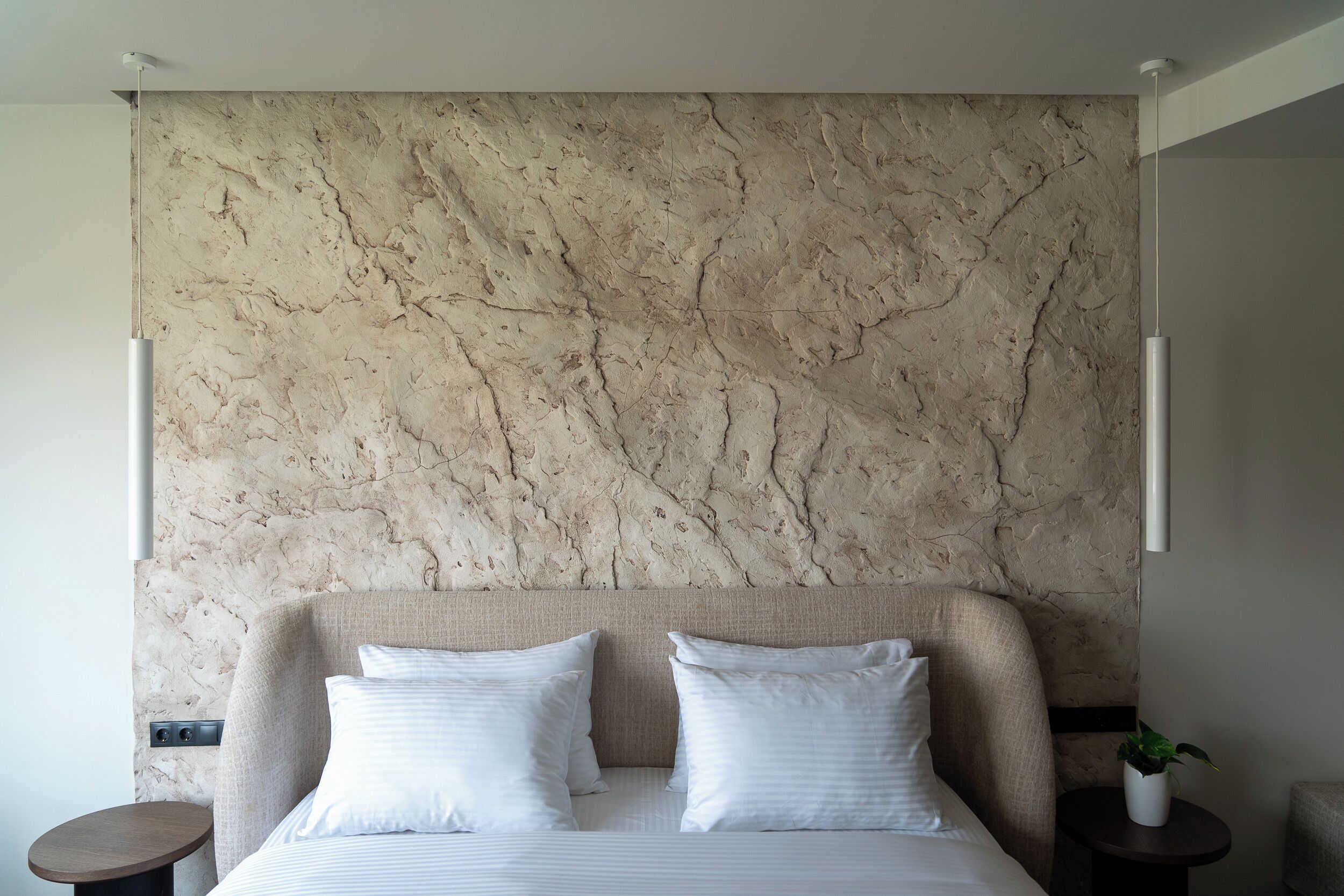 Habitación de hotel moderna con cabecera de mármol, cama bien hecha con sábanas blancas y una pequeña mesa redonda con una planta.
