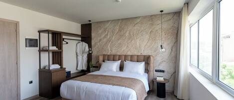 A modern hotel room with a large bed, a wardrobe, a vanity with a mirror, and a window with a view.